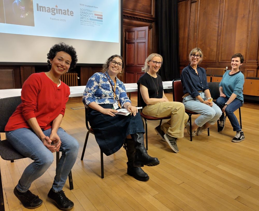 Five cheerful women sitting on chairs in front of a large projection screen, in a traditional panelled room