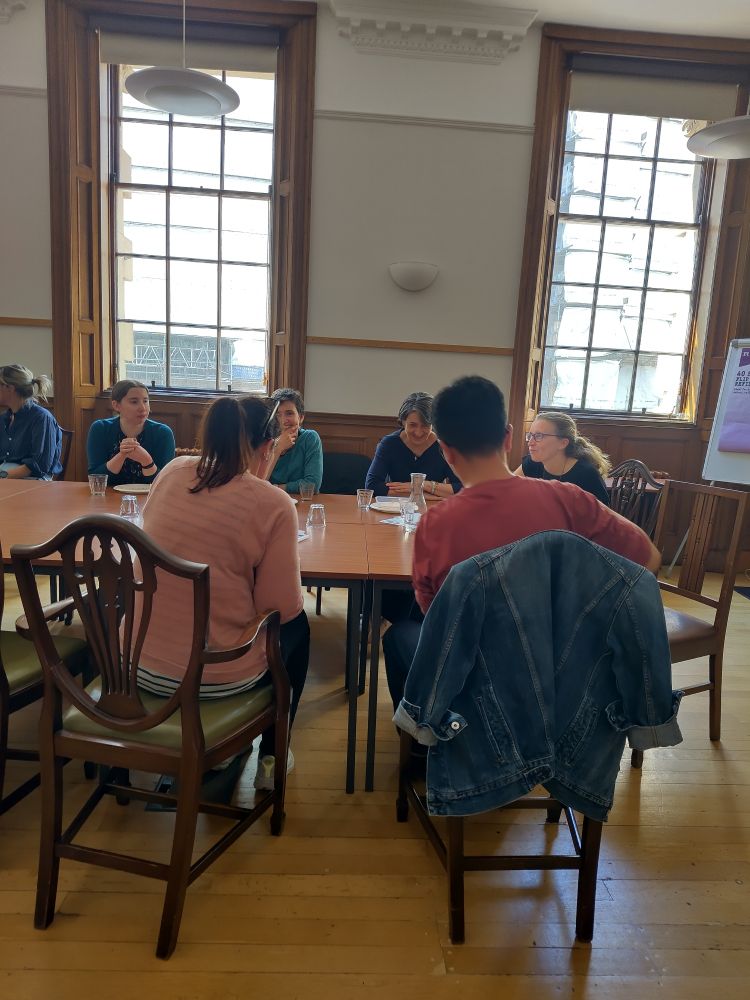 Several people sitting at a long table engaged in conversation inside a large room with tall windows 