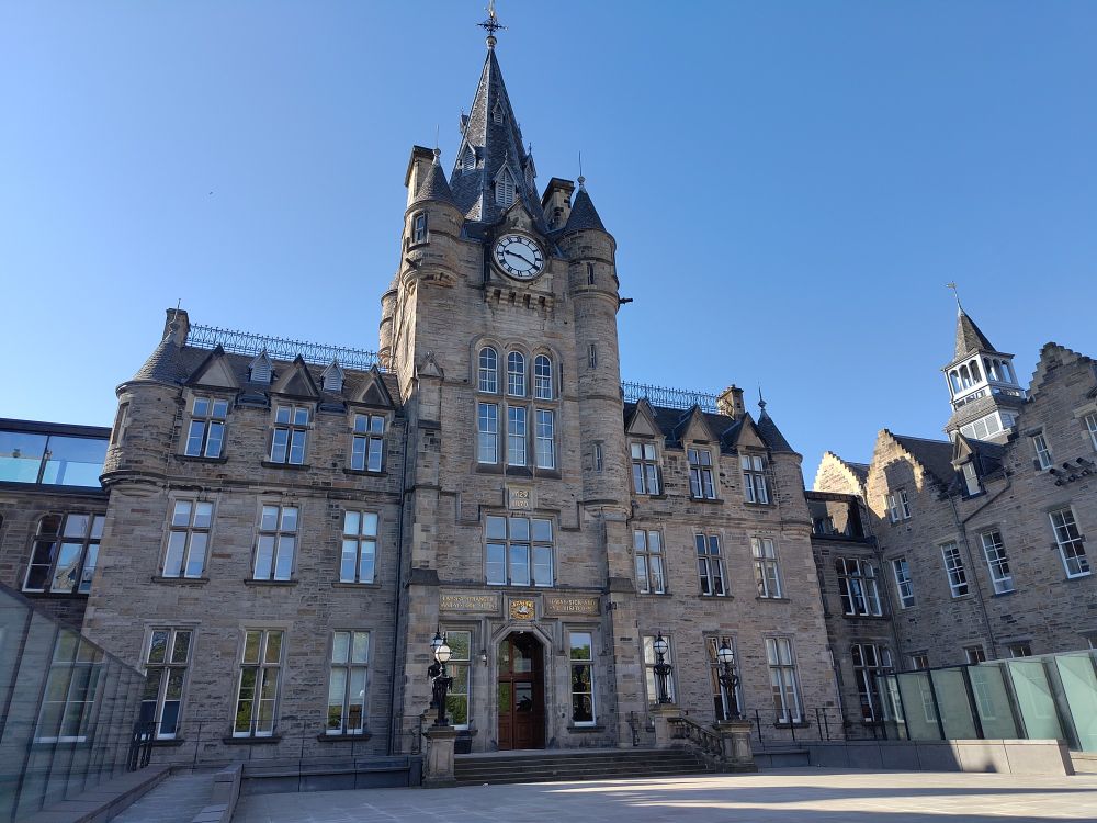 Imposing and beautiful stone built Edinburgh Futures Institute, with taller centre section with conical roof above, clock near the top, and a wing each side with 9 windows on each, pointed dormer windows across the tops
