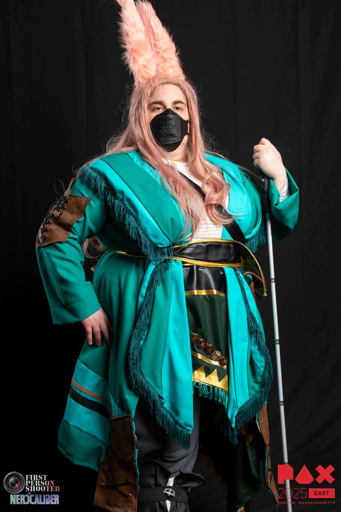 A photograph of myself in Taapu from Final Fantasy 14 cosplay, a rabbit eared character with a teal coat with many leather details, a black and gold leather belt, several leather pouches, a twisted teal and aqua scarf with dark green fringe, and a second scarf around the waist with golden beads and fringe. I am carrying my blind cane, as both myself and Taapu are blind.