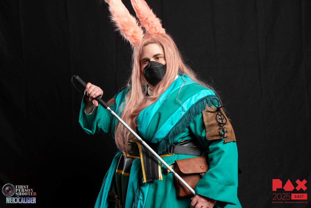 A photograph of myself in Taapu from Final Fantasy 14 cosplay, a rabbit eared character with a teal coat with many leather details, a black and gold leather belt, several leather pouches, a twisted teal and aqua scarf with dark green fringe, and a second scarf around the waist with golden beads and fringe. I am carrying my blind cane, as both myself and Taapu are blind.