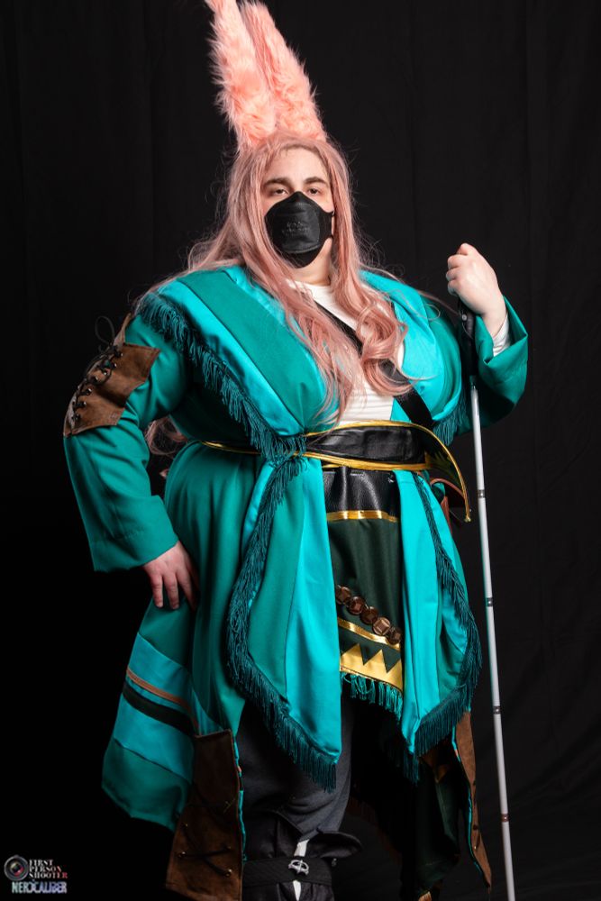 A photograph of a cosplay of Taapu from Final Fantasy 14, in the Dawntrail expansion. I am dressed as her, in a knee-length open green coat made of a loose woven teal fabric, with a series of horizontial stripes near the lower hem in light teal, tan, and dark green. I am wearing a fringed scarf, rippling with light and dark teal fabric around my shoulders going down. The jacket also is belted, with the scarf belted in as well with a black leather belt with gold accents. The jacket also has a number of leather details, fastened with metal eyelets with dark leather cord. I am wearing a white shirt and heather grey pants beneith, though most is hidden by the jacket and scarf. A second, shorter scarf is belted around my midsection with fake metal coins, gold detail, and teal fringe. I am wearing a shoulder length pink wig with pink furred rabit ears, wearing a KN95 mask, and holding my white blind cane. In the bottom left corner is the photographer's logo for Nerd Caliber.