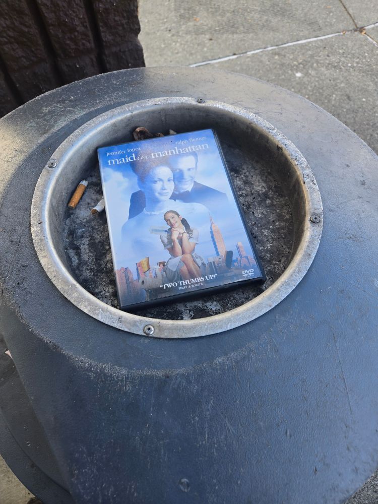 a copy of the movie "maid in manhattan" with Jennifer Lopez and some guy on dvd sitting in the ashtray part of a public garbage can outside in natural daylight. the DVD is surrounded by cigarette butts