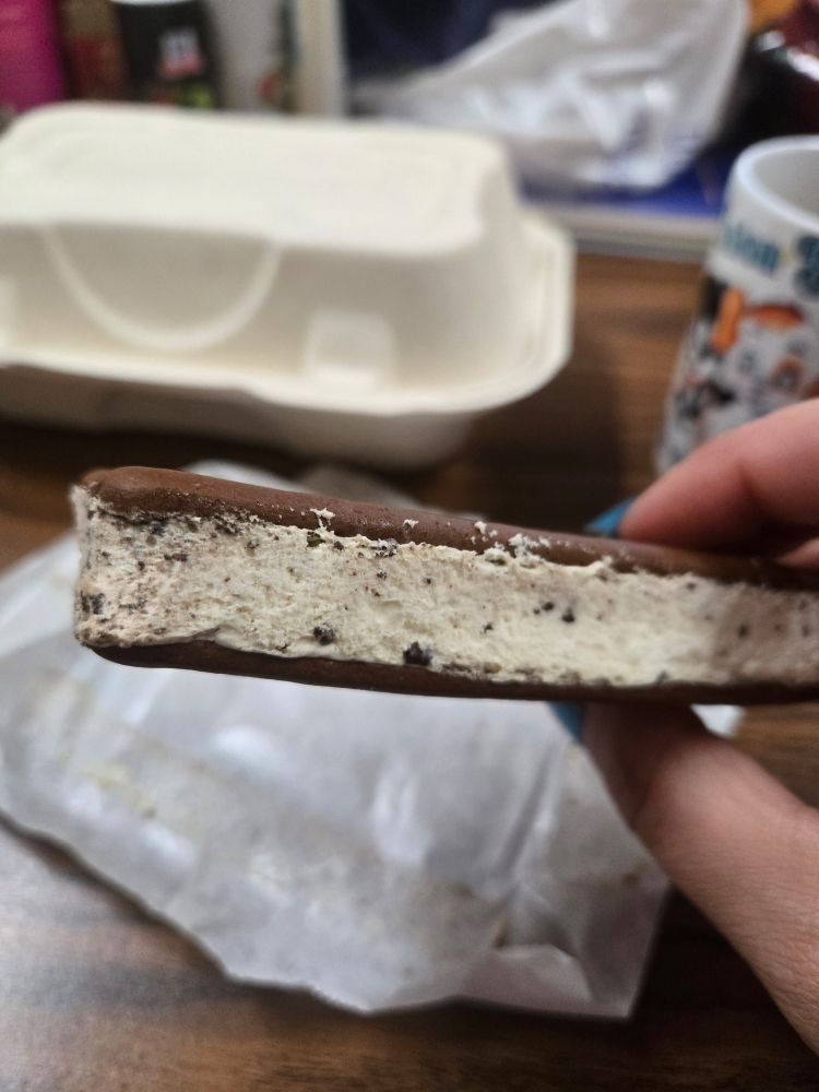 my hand holding a cookies and cream ice cream sandwich over a cluttered wooden table with the wrapper in the background as well as a closed (and empty) fast food container