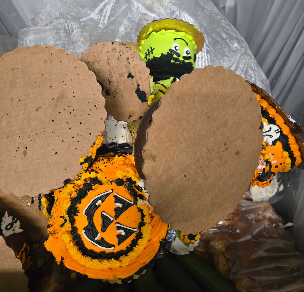 A bunch of grocery bakery cakes decorated with orange and black pumpkins and green Frankenstein's monster on cake plates dumped unceremoniously into a compost bin