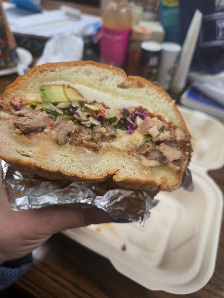 my hand holding the second half of a carnitas torta over a cardboard takeout package sitting on a cluttered wooden table where many people gather and eat daily under florescent lighting. interior shot