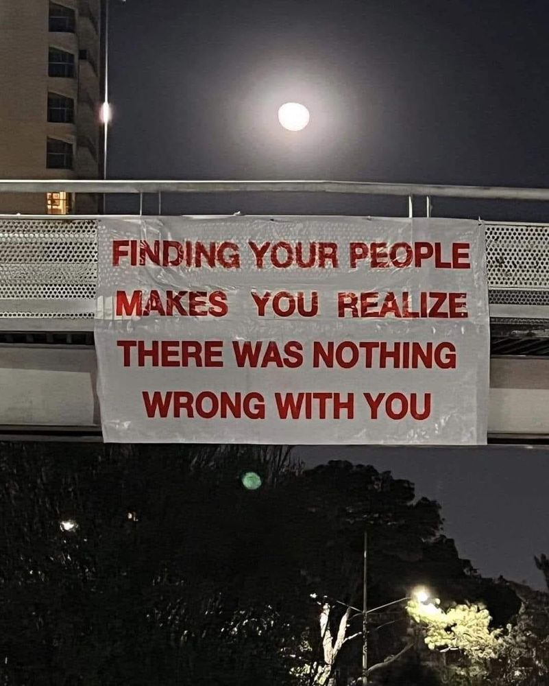 a photo of a white poster hanging from a bridge, reading in red letters: "finding your people makes you realize there was nothing wrong with you", taken at night with moonlight shining from above