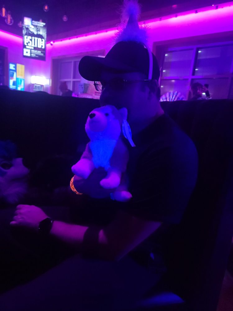 Picture of Oddy at a nightclub with a plush dog in his hand and a birthday hat on his head.