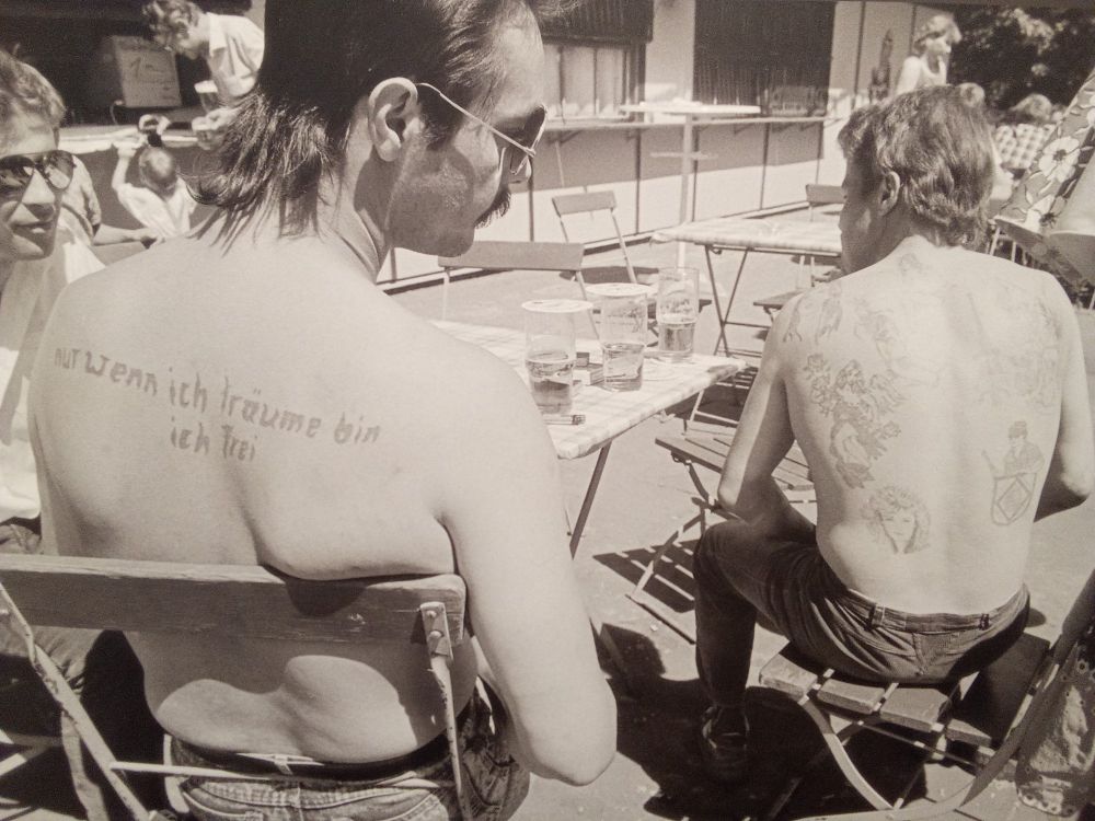 Photo avec deux hommes torse nu. Celui de gauche à un tatouage avec écrit "nur wenn ich träume bin ich frei"