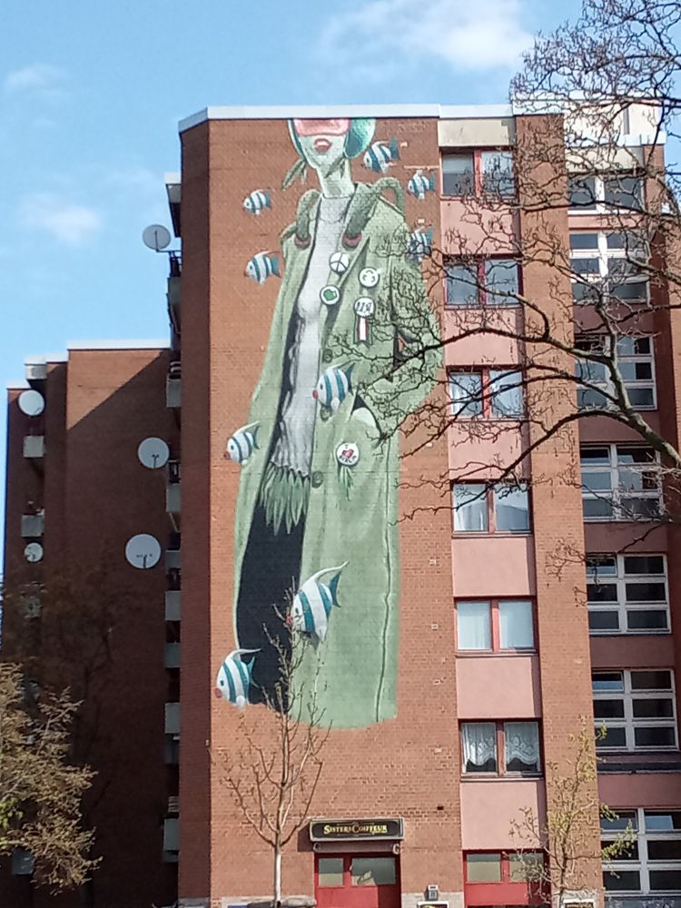 Graff sur toute la hauteur d'un immeuble berlinois représentant le corps d'une femme dans une veste verte entourée de poissons bleus et blancs 