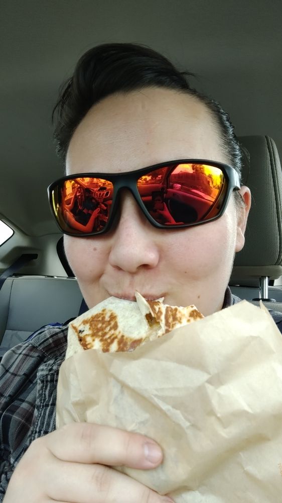 Selfie of Jess in a car wearing red tinted UV sunglasses while taking a bite out of a breakfast crunchwrap from Taco Bell 