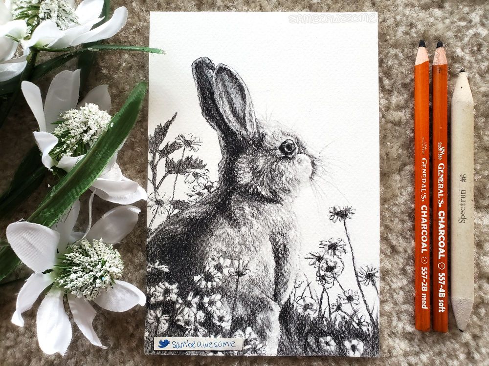 A photo of a charcoal drawing of a rabbit that's surrounded by flowers and grass. Faux flowers are next to the artwork for flair, along with the pencils used to make the artwork.
