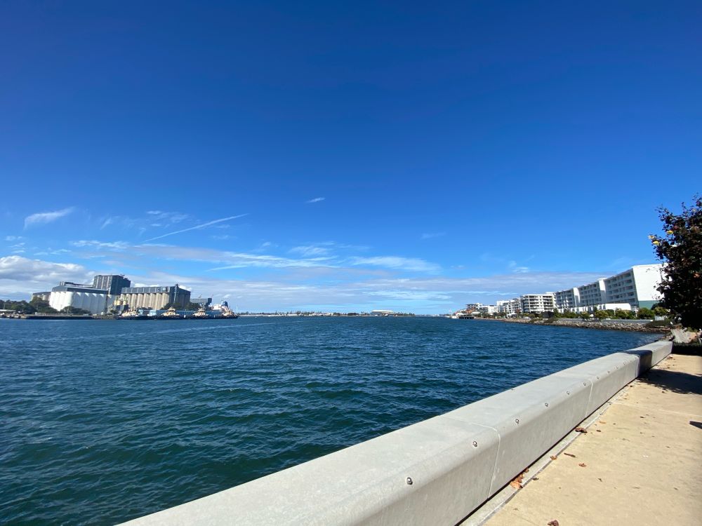 Photo of a harbour, calm water looking out into the ocean. There are some industrial buildings on the other side of the bay, and a footpath stretching into the distance on this side. 
