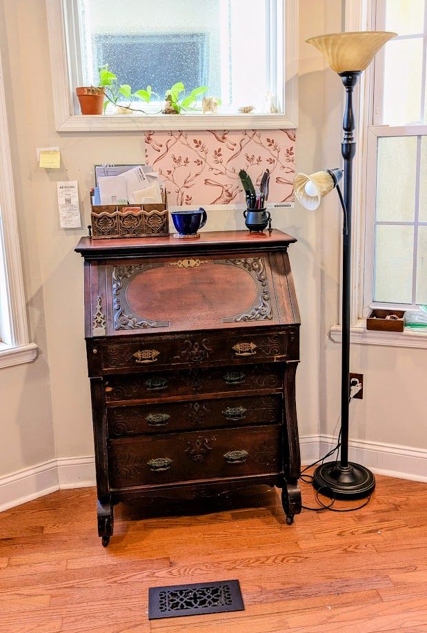 Photo of a wood secretary desk with four large drawers up against a wall. The desk has carved detailling and decorative metal drawer pulls. Sitting on top of it is a paper organizer, a large blue mug, and a small cauldron with pens and quills in it. Beside the desk is a standing lamp, and above it is a houseplant on a windowsill.