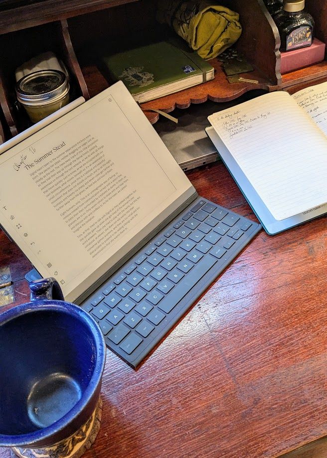 Photo of a desk with a large empty mug, an open tablet on a page full of text titled "Chapter 16: The Summer Stead," and an open notebook with handwriting. In the desk's shelves are more notebooks, a jar, bottles of ink, and a fabric roll of pens. 