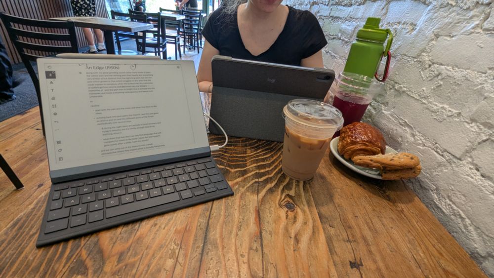 A photo of a table at a café with an iced chai latte, a hibiscus iced tea, a cookie, and a chocolate croissant. Sitting across the table is a person (face not visible) typing on an iPad.