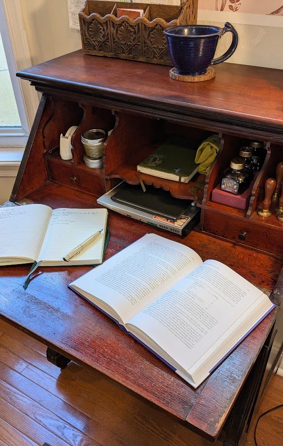 The same desk, open, with a book and a notebook laying open on its surface. The notebook has an uncapped fountain pen resting on it, and on the shelves of the desk there is a tape dispenser, a jar of green salve, a laptop, a notebook, bottles of ink, and several wax seal stamps.