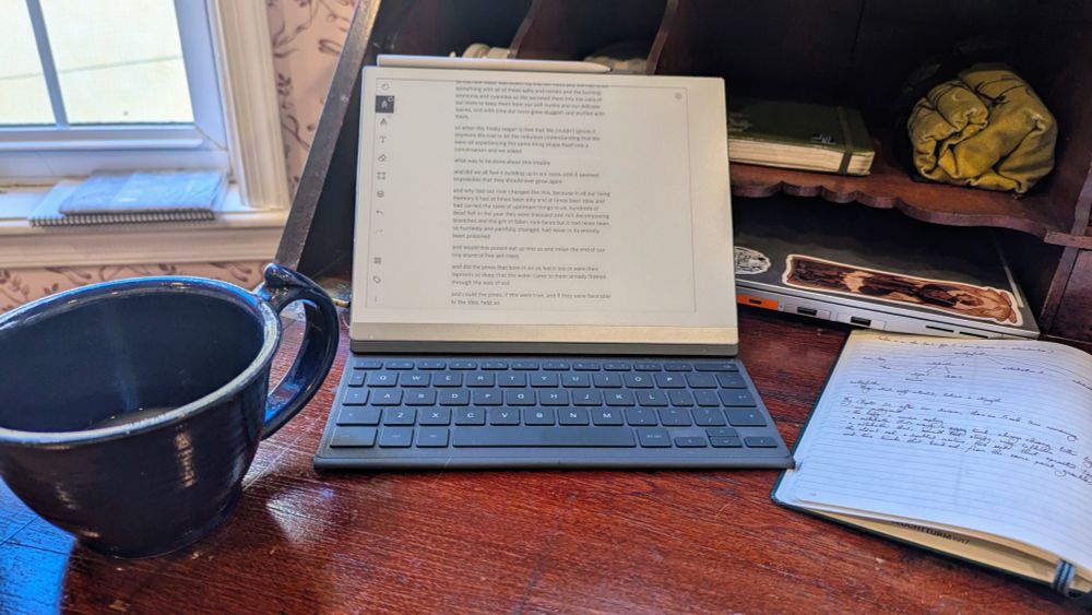 Photo of a tablet on a reddish wood desk, with an empty blue ceramic mug to one side and a notebook to the other. In the desk shelves a laptop is visible, as are a roll of pencils and another notebook.