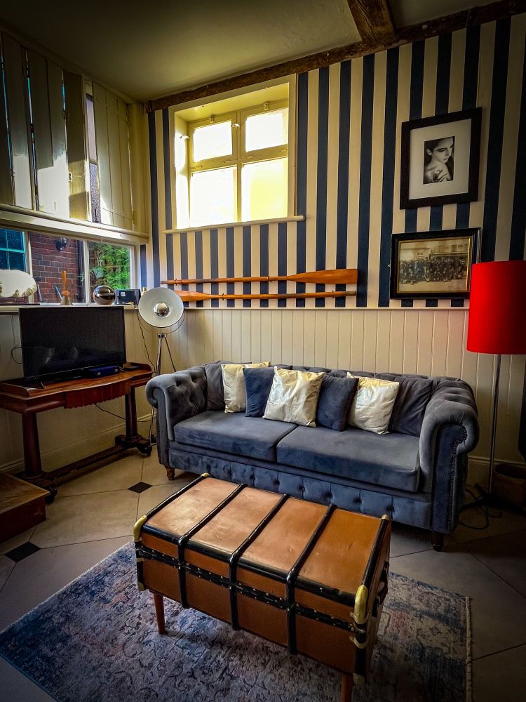 Ein kleiner, stilvoll eingerichteter Raum mit einer dunkelgrauen Samtcouch, die mit glänzenden Kissen dekoriert ist. Davor steht ein Koffer, der als Couchtisch dient, auf einem Teppich. Die Wand ist blau-weiß gestreift, geschmückt mit Bildern und zwei hölzernen Rudern. Eine rote Stehlampe setzt einen kräftigen Akzent, während ein kleines Fenster warmes Licht hereinlässt – eine Couchgeschichte zwischen Vintage-Charme und maritimen Details.