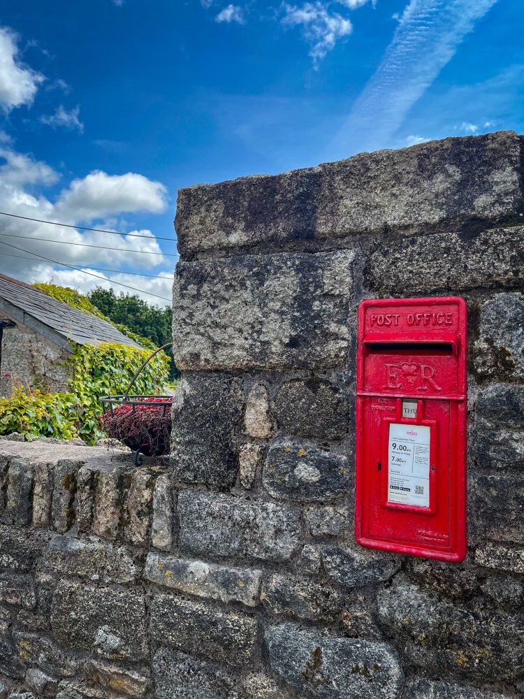 Ein roter Briefkasten der britischen Post ist in eine alte Steinmauer eingelassen. Darüber steht „POST OFFICE“ und das königliche Monogramm „E II R“. Der Himmel ist blau mit einigen weißen Wolken, und im Hintergrund sind ein Hausdach, grüne Pflanzen und Stromleitungen zu sehen.