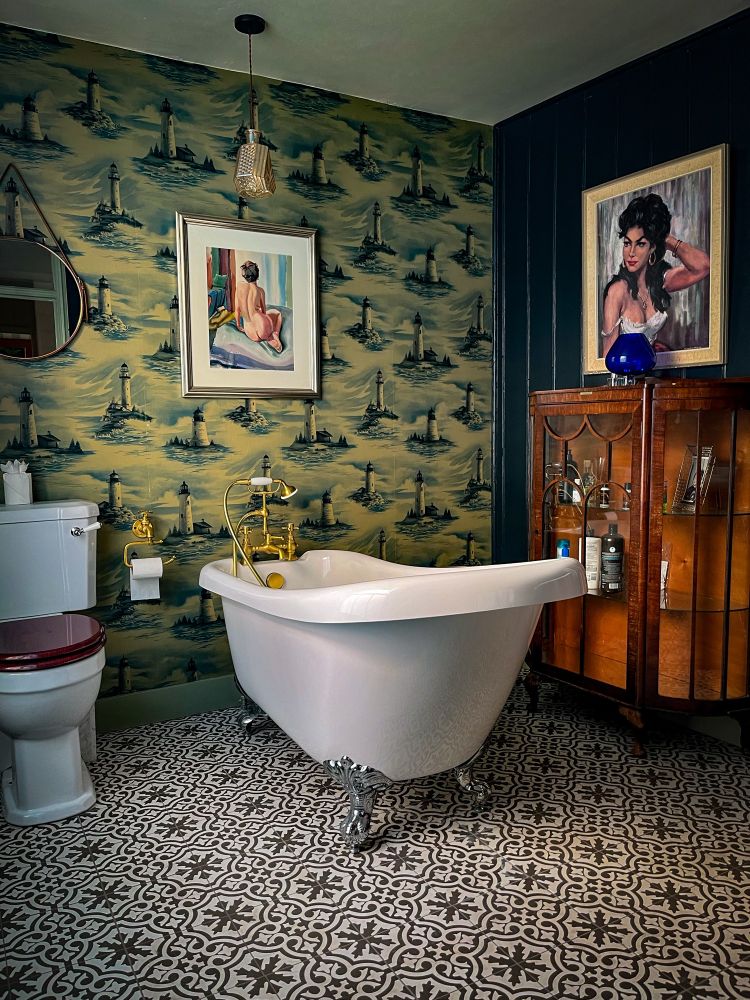 Ein stilvoll eingerichtetes Badezimmer mit einer freistehenden weißen Badewanne auf dekorativen Löwenfüßen. Die Wand hinter der Wanne ist mit einer Tapete im Leuchtturm-Motiv bedeckt. Links neben der Wanne stehen eine Toilette und ein kleiner runder Spiegel. Über der Toilette hängt ein gerahmtes Gemälde einer sitzenden Frau von hinten. Rechts der Wanne steht eine antike Vitrine mit Glasfront, darauf ein blaues Glasobjekt und darüber ein weiteres Porträt einer Frau. Der Boden ist mit schwarz-weiß gemusterten Fliesen ausgelegt, und eine einzelne Hängelampe mit goldenem Schirm hängt von der Decke.