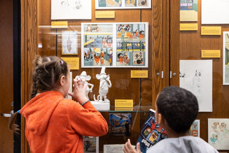 Young children (one white, one black) looking at Team Pom display of scale model of Mr. Gilbert and Monsieur Georges (rats in suits) made of white polymer clay. Digital printouts of a subway chase scene from Team Pom Volume 2: The Last Dodo in the background.