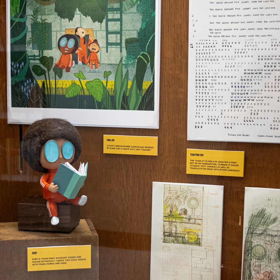 Handwriting specimen for conversion into typeface. Scale model of Ruby (young black girl with glasses, afro and wearing a red-orange tracksuit) made of wool, fabric and clay. Background contains artwork from Team Pom volume 2: The Last Dodo, as well as sketches.