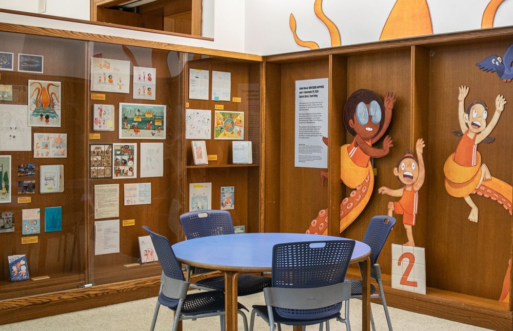Exhibition of work-in-progress sketches of The Adventures of Team Pom at the Youth Wing of Central Brooklyn Public Library. Display of characters—Ruby, black girl with glasses and an afro, Roberta a small asian character with a top knot, and Agnes, a Mexican American girl with pigtails. Vitrines contain sketches, several versions of Squid Happens covers.