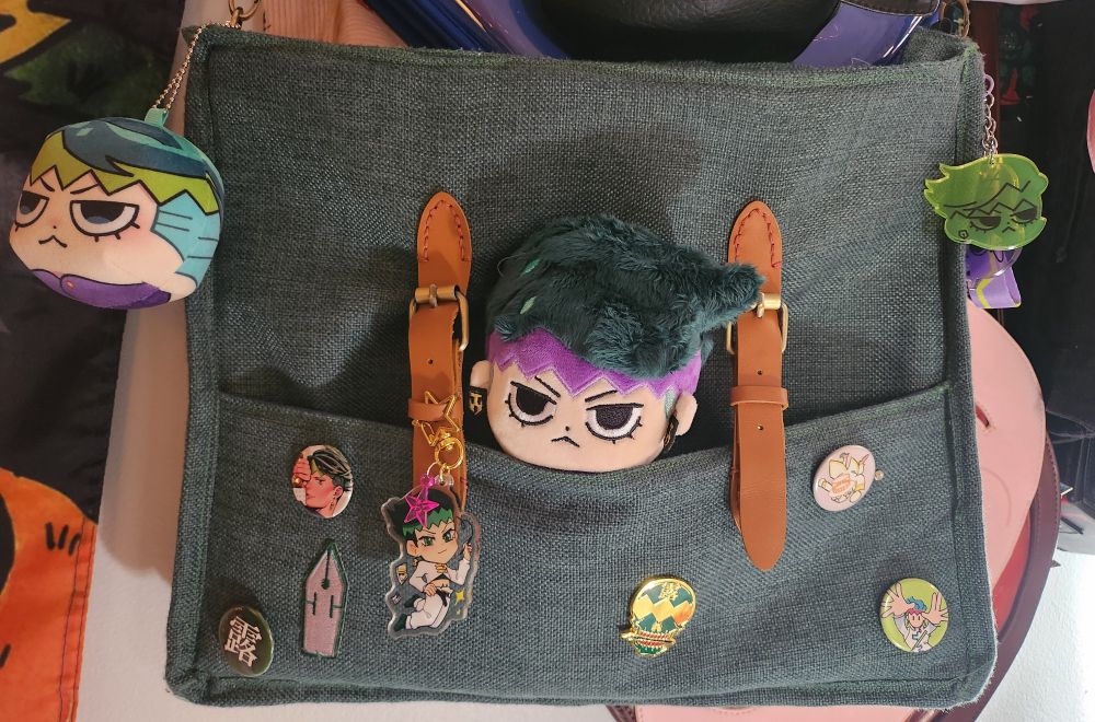 A Rohan Kishibe themed bag with five pins, three keychains, and one plush designed after him. The bag itself is green and has two brown straps, and is designed after a sketch book the character carries.