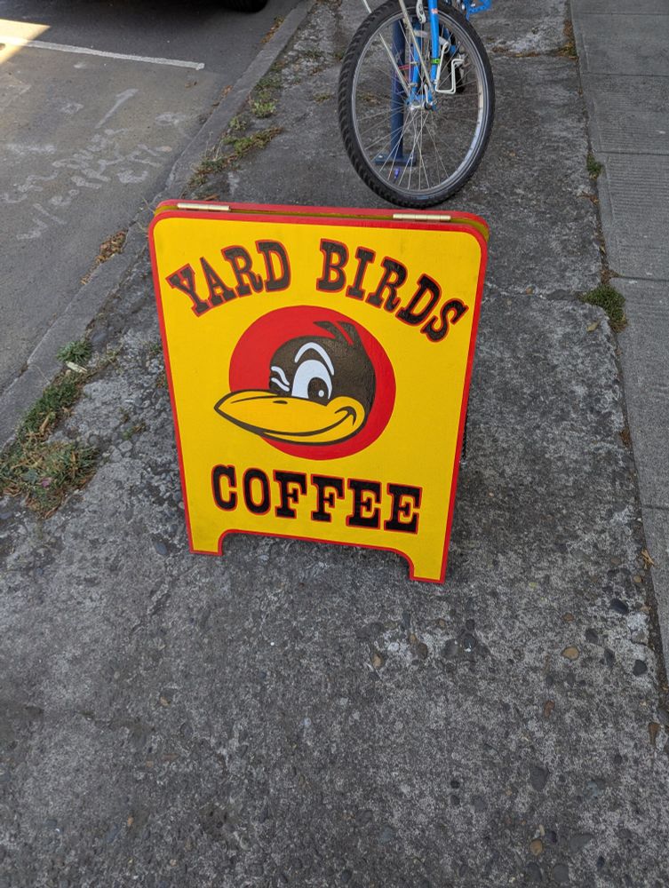 A sign on the sidewalk at 42nd and Prescott in PDX. Vibrant Yellow and red advertising "YARD BIRDS COFFEE", with the iconic black yard bird winking.

A bicycle, parked nearby.