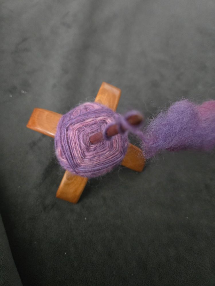 Wooden Turkish Drop Spindle. The spindle is a bottom cross whorl, with purple yarn wrapped around the arms of the "cross."