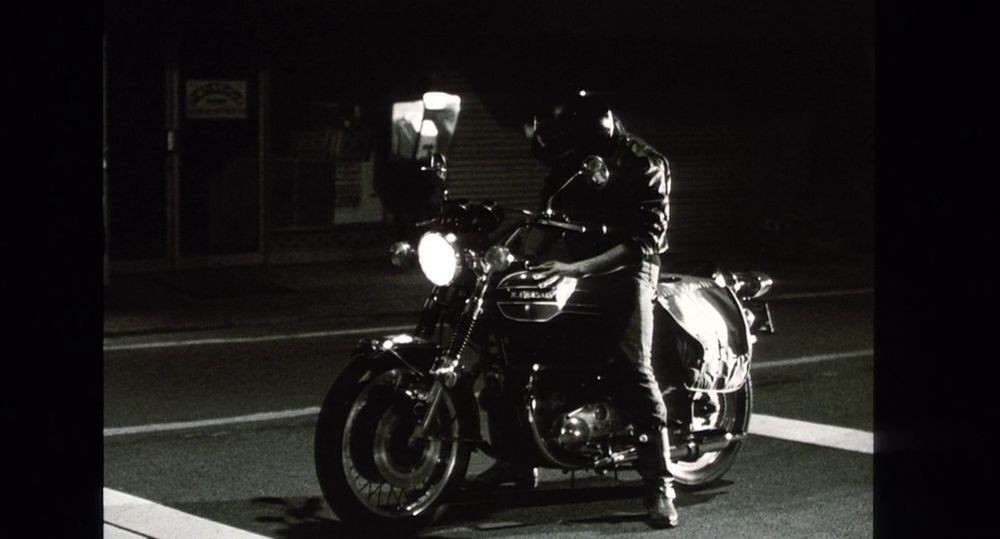 still from His Motorbike, Her Island. black and white. a man in biker attire is caressing his motorbike