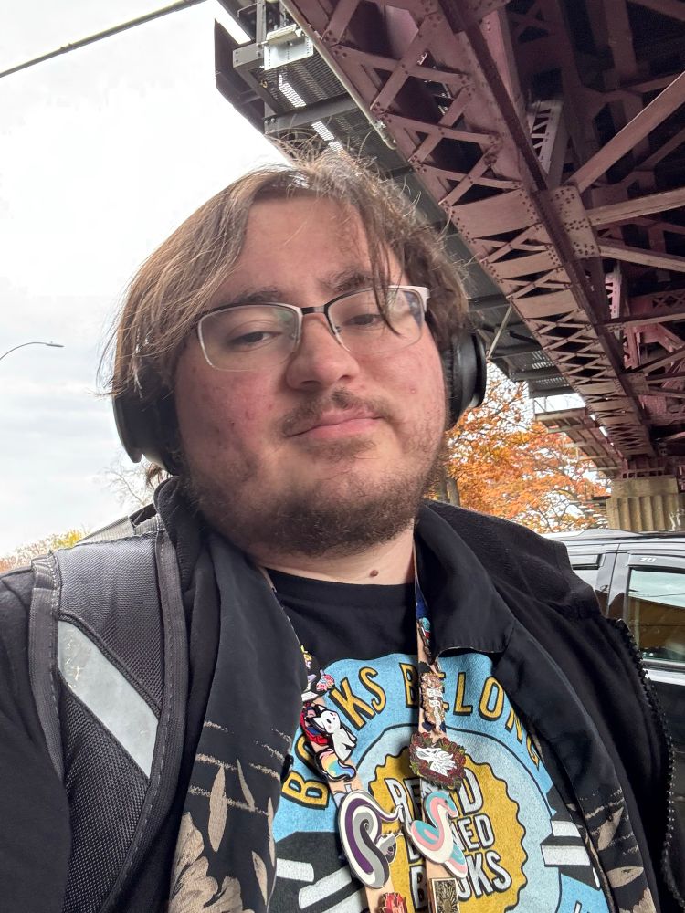 Sam with a lanyard with too many pins, a read banned books shirt, and headphones