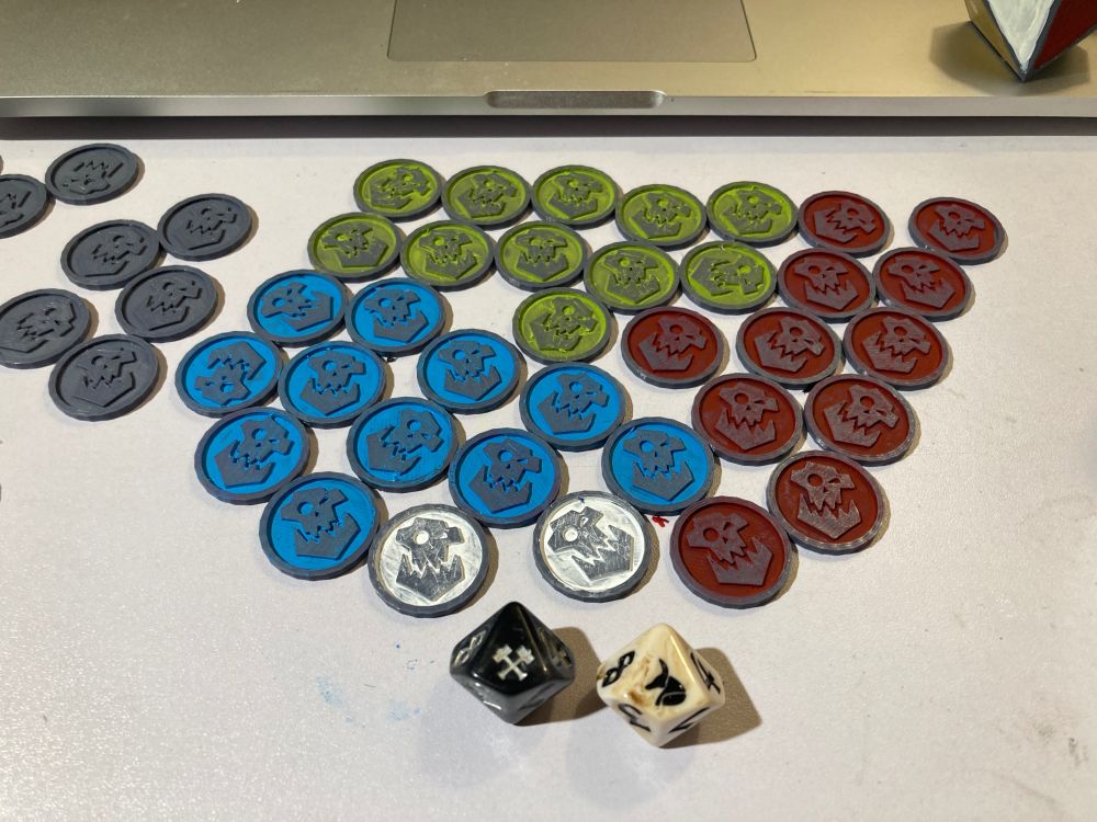 a bunch of flat orc tokens with Warhammer-esque iconography. Each is a round 28mm disk with a large-jawed orc face icon on it. I've painted the backgrounds blue, slime green, blood red, or white, leaving the orc face icon unpainted.
In the foreground is a black d10 with silver crossed hammers on the top face and a bone-desert-colored d10 with Anubis' head in profile as the top face.