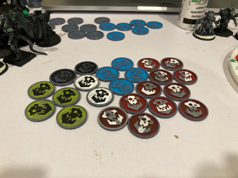 A bunch of flat-ish 3D printed tokens with abstract orc faces. Black with green background, black with white background, grey with blue background, grey and white with deep red background. 