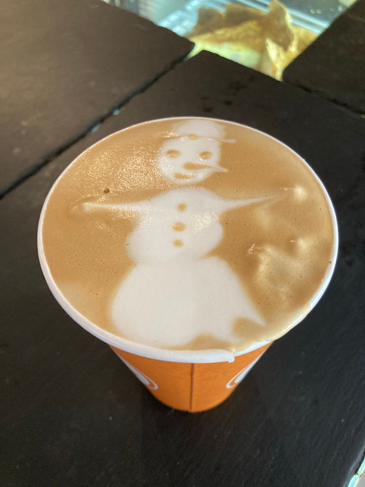 Paper coffee cup, latte art of a snow person with a top hat and their arms outstretched for a hug