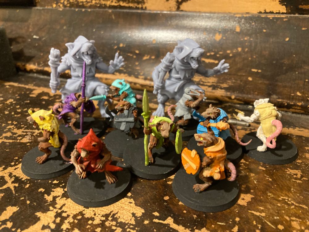 Photo of my painted ratmen minis standing around two (grey) unpainted ratmen minis that are twice as tall as the painted ones.