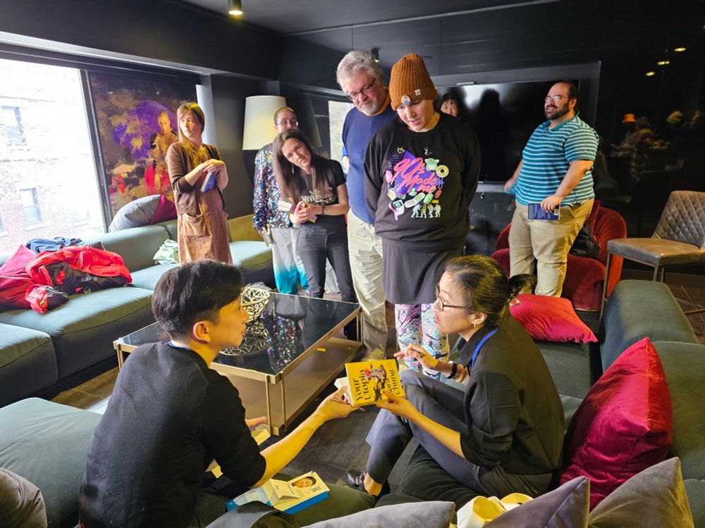 Author Bora Chung and translator and author Anton Hur signing a copy of her short story collection "Your Utopia". a few people are lining up in the background of the picture, which was taken in a hotel suite furnished with couches, chairs and a low table.
