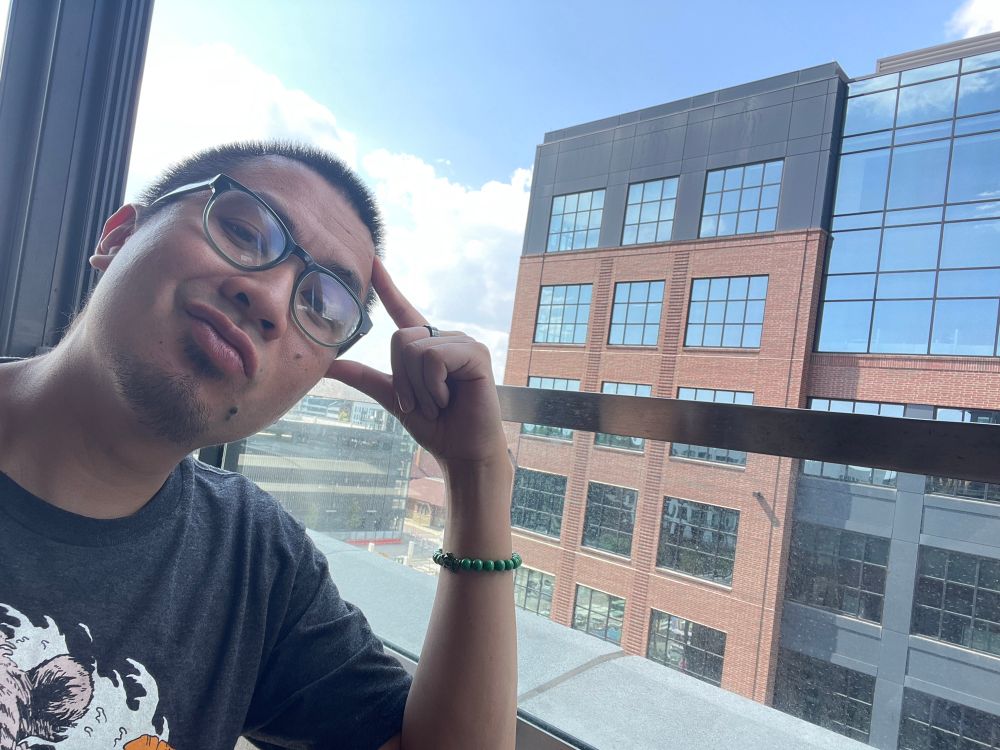 An Asian American dude with glasses sits at a rooftop bar, with sunny blue skies and a black-brown building behind him.