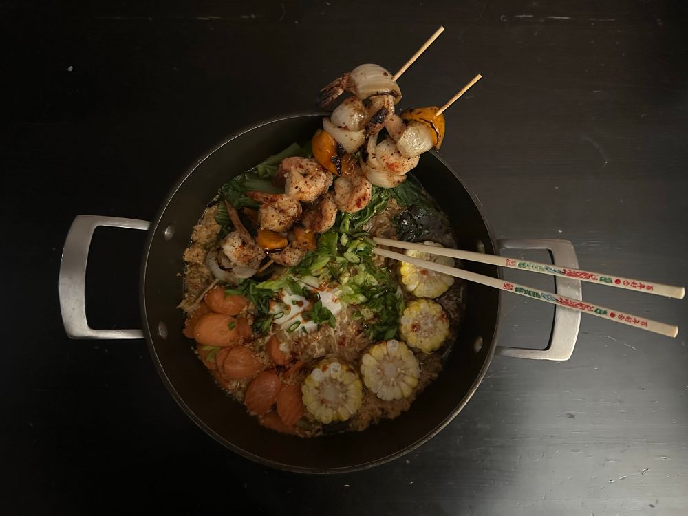 Ramen with shrimp skewers, grilled corn, bok choy, and sliced carrots with a green onion garnish.