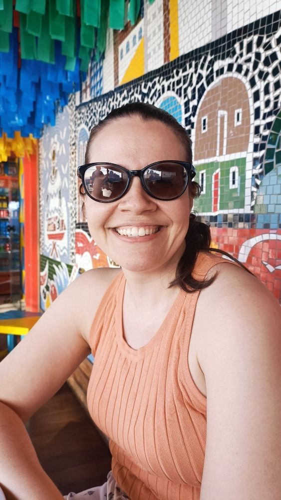 Foto minha sorrindo. Tem uma parede de mosaicos coloridos atrás de mim.