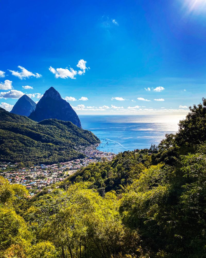 The Gros and Petit Piton are volcanic plugs in Saint Lucia, part of the Soufrière Volcanic Center. Picture is from above in the mountains overlooking the town of Soufrière nestled into a valley that sits on the coast. Gorgeous blue waters, boats and pointy volcanic mountains dot the coast. 