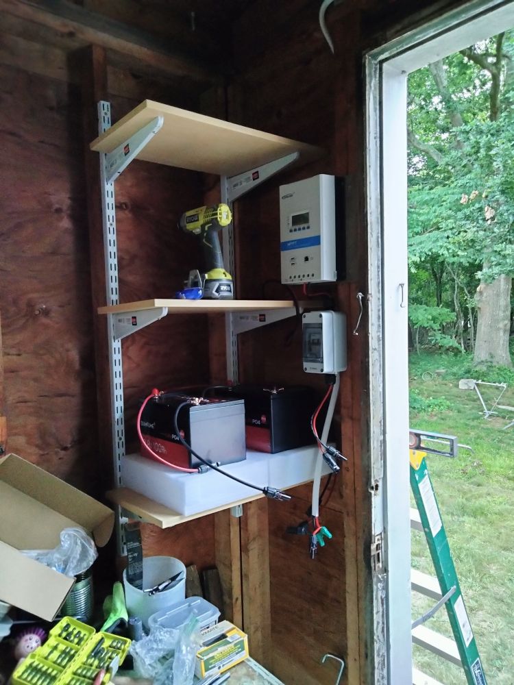 The corner of the back room of our island cottage, with some freshly installed shelving.  We have two lithium iron phosphate batteries on the bottom shelf, connected to the solar panels, with regulators and other devices about which I do not yet have a clue. 