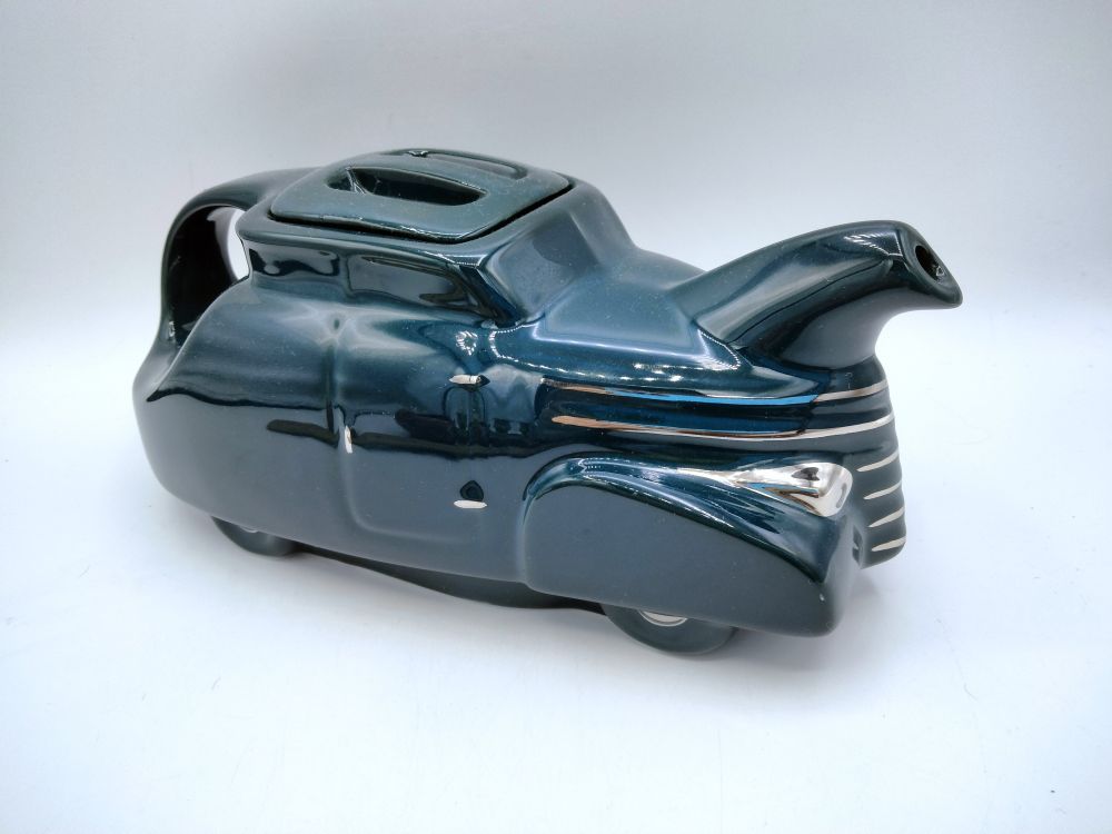 A dark teal automobile shaped teapot from China Specialties.  This is a reproduction of Hall China's Automobile teapot.  I am not sure what kind of car it is supposed to represent, but it does look to be a 40s style.  The handle is in the back, a wide band going from rim to "trunk".  The spout is on top of the hood, over a grille accented with silver/platinum bands.  The lid is square with rounded corners, and indented to form a bar handle in the middle.  
