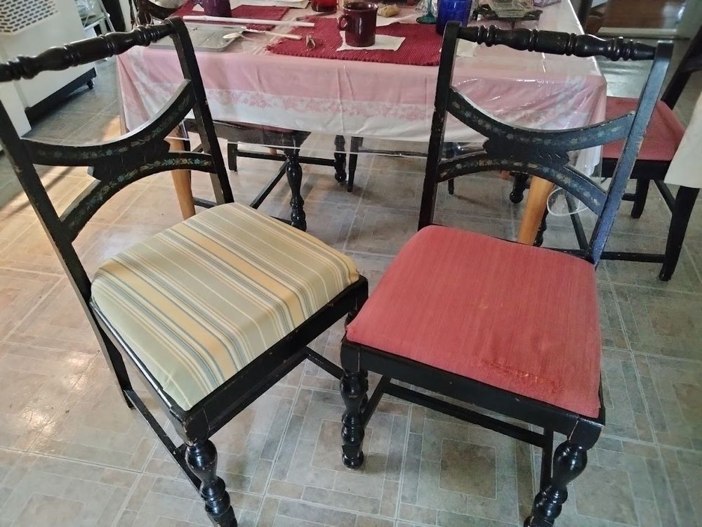 Before and after photos of vintage kitchen dining table chairs on which I redid the seats.  They are from the 20s according to a date I found on the bottom of one! The backs have curved side pieces, a turned spindle type top bar, and a neat double arc middle piece, with an upward curve on top and a downward curve below. The front legs are also spindle/turned, and there is an H support connecting all four legs. I purchased them with a reddish, faintly striated cloth covered pad which was thin and worn (chair on the right), and replaced that with a thicker foam and a pale blue, yellow and green fabric which echoes the colors of the likely home-stenciled flower garland decoration across those arcs on the backs (chair on the left). So comfortable! 
