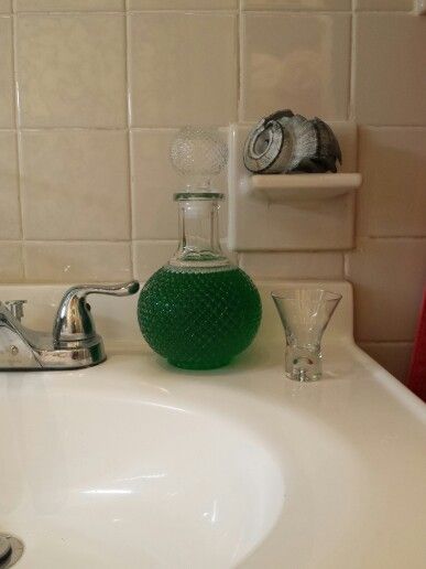 uma garrafa de vidro com tampa tbm de vidro cheia com  enxaguante bucal (a cor do liquido é verde escuro), a garrafa tem textura de varios prismas parecendo escamas bem pequenas, ela tem o formato classico de poção magica, bem redonda em baixo, a tampa é igual a garrafa 