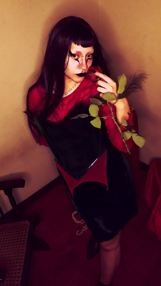 em pé uma garota, magra e branca (eu) com cabelos escuros franja vegana, batom preto e delineador comprido tanto pra cima quanto pra baixo do olho, segurando uma rosa perto do rosto usando uma saia com uma ponta assimétrica maior no lado direito em veludo preto, segunda pele vermelha com duas pontas no centro e na altura do cotovelo tem argolas de metal, corset de vinil, bota bico fino preta. 
a foto tem um ângulo de cima pra baixo, e cor quente meio filtro america latina 