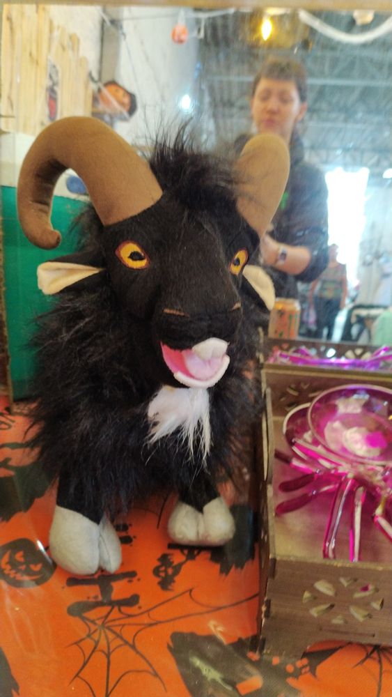 A mais perfeita pelúcia de bode preto com cavanhaque branco, ele tem pelos compridos pelo corpo todo menos na cara patas e chifres, olho amarelo típico de bode, no fundo da imagem tem um espelho sujo e ta refletindo uma coitada (eu)