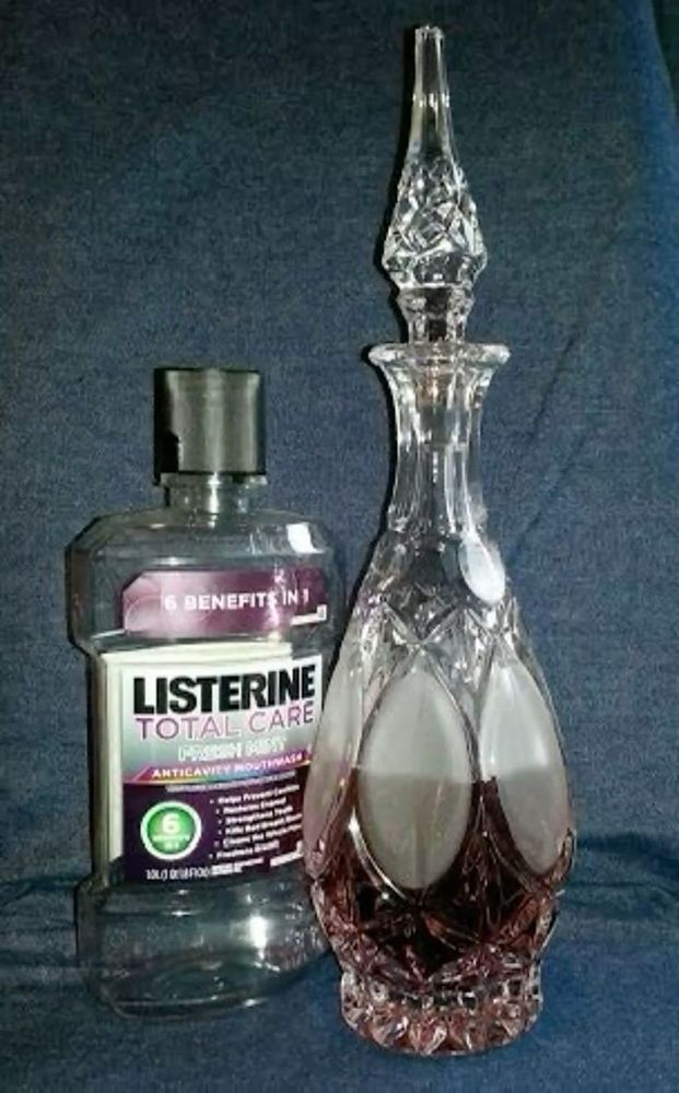 garrafa de listerine total care vazia do lado de uma garrafa de vidro com tampa tbm de vidro cheia com o listerine (a cor do liquido é lilás), a garrafa tem mtos detalhes e texturas, o formato da garrafa é em gota e a tampa é quase um prisma 