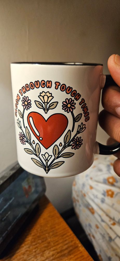 White mug with a black handle with the phrase "Bloom Through Tough Times" above a red heart surrounded by flowers on the front.